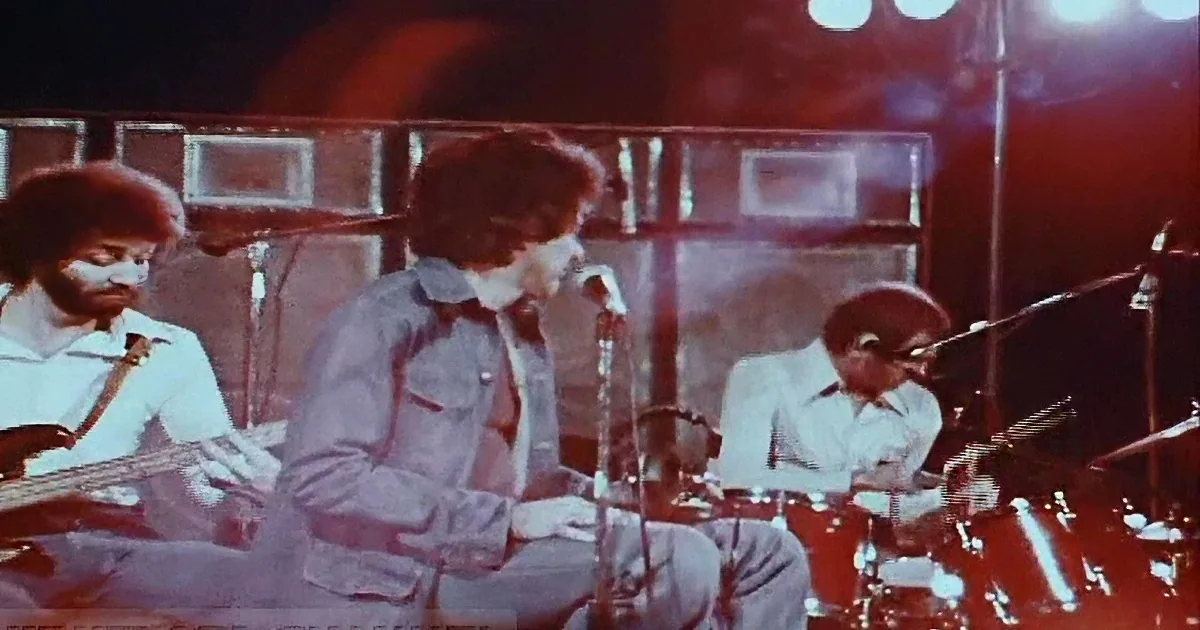 A band performs on stage with a guitarist, bassist, and drummer under bright lights.