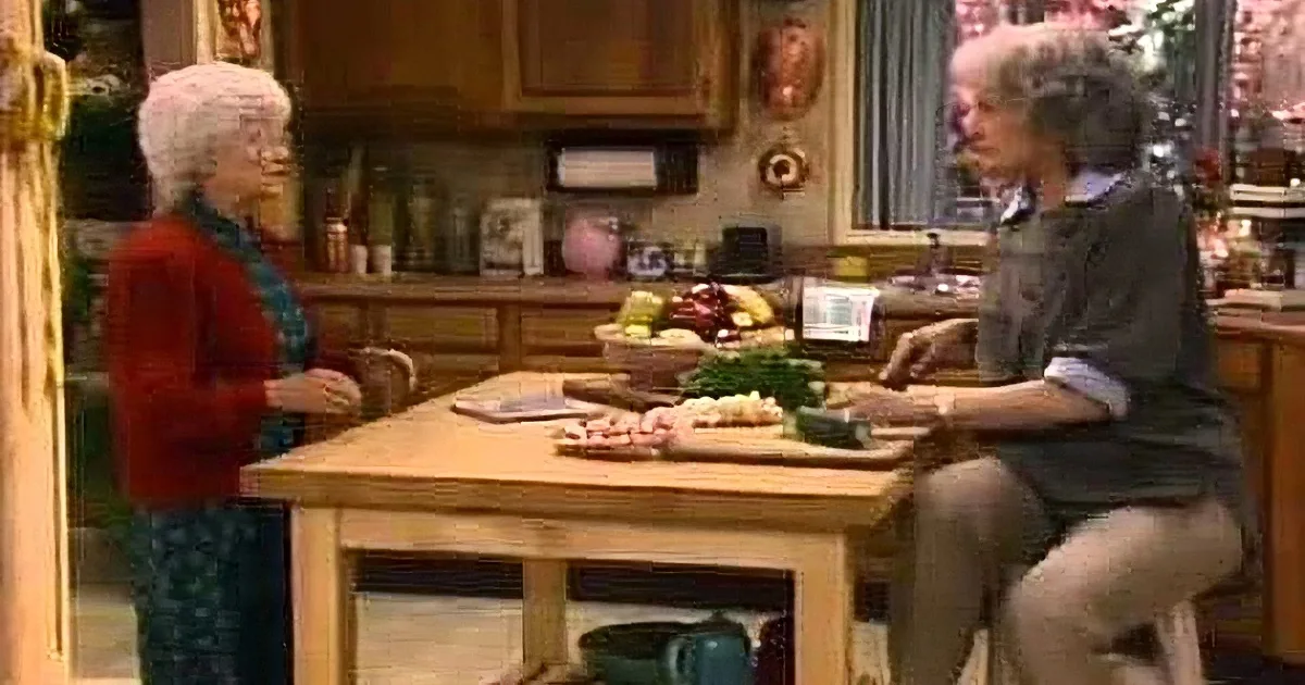 Two women in a kitchen, one standing at a table with food, the other observing.