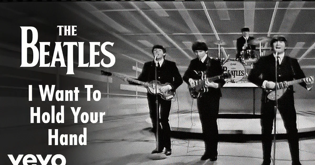 The Beatles perform on stage in black suits, holding guitars, with a drummer in the background.