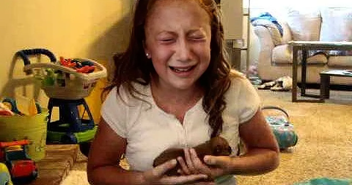 A young girl is sitting on the floor, holding a toy, with a distressed expression on her face.
