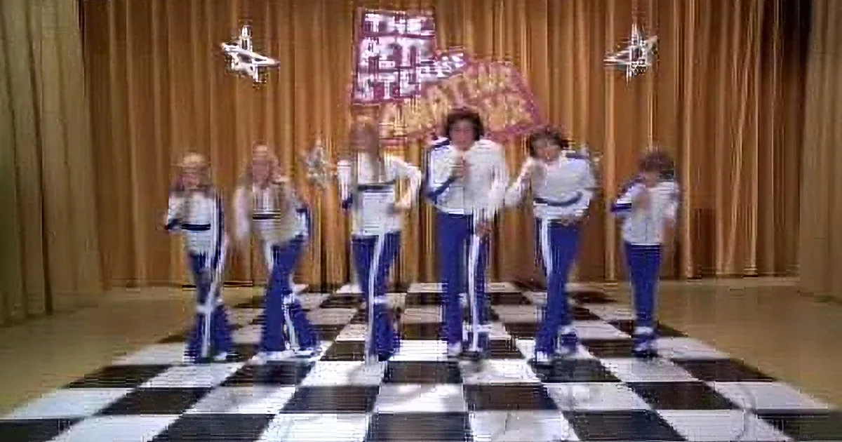 A group of performers in blue and white outfits dance on a checkered floor, with a backdrop featuring "Pete Steiner" and...