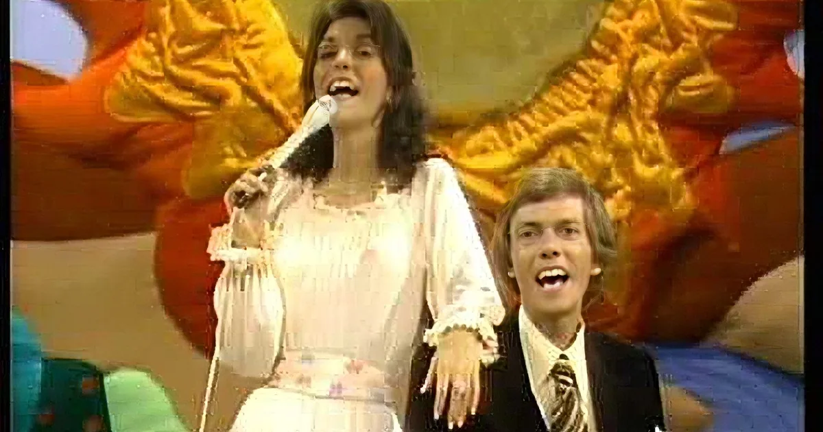 A woman in a white dress sings into a microphone, surrounded by colorful, vibrant stage decorations.
