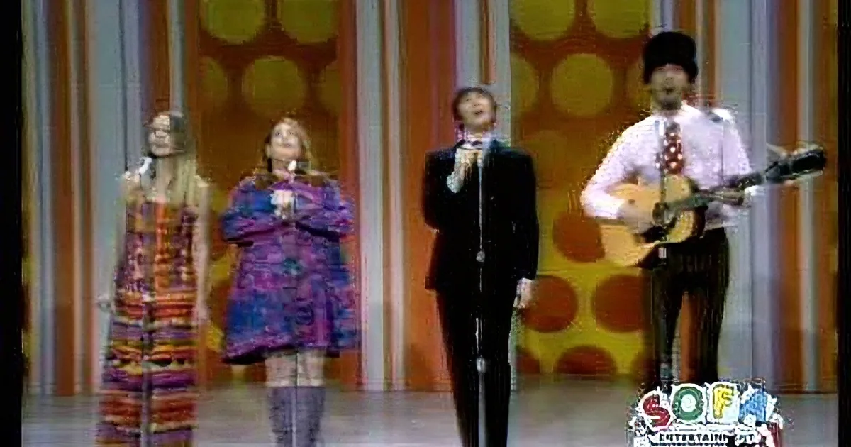 A group of performers in colorful outfits sing and play guitar on a stage with a vibrant background.