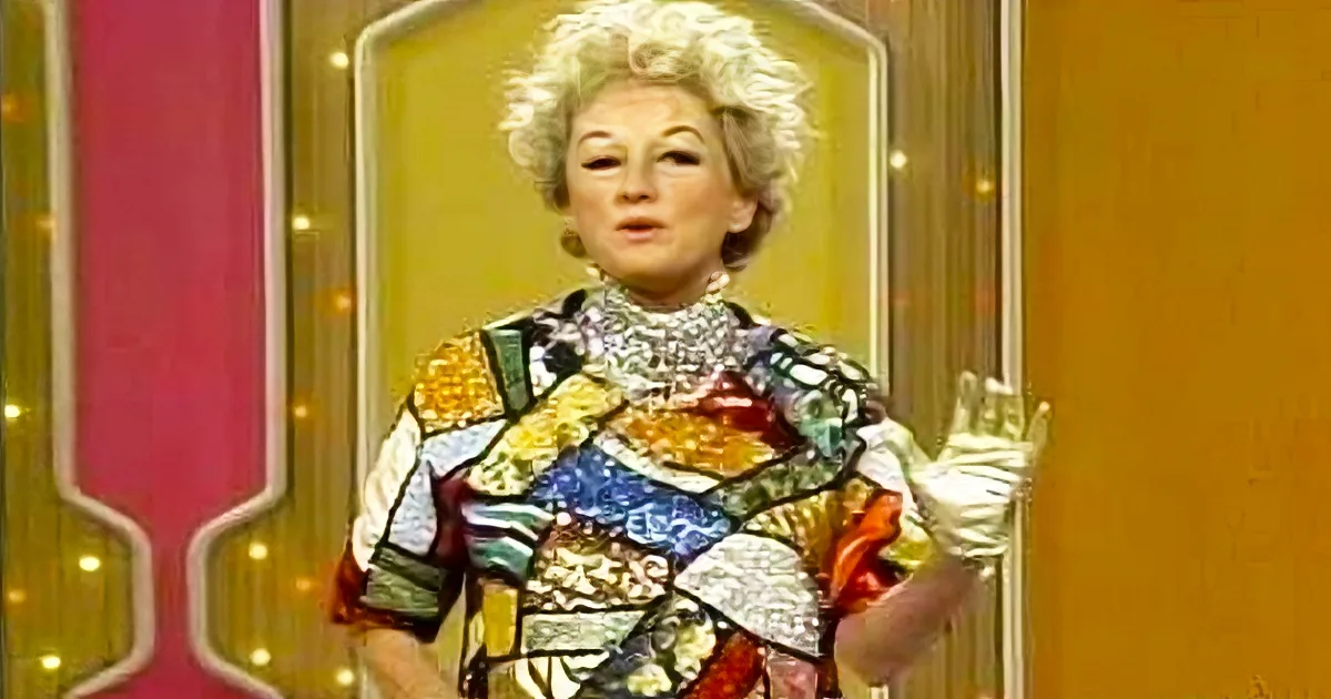 A woman in a colorful, patterned outfit stands in front of a decorative backdrop, gesturing with her hands.