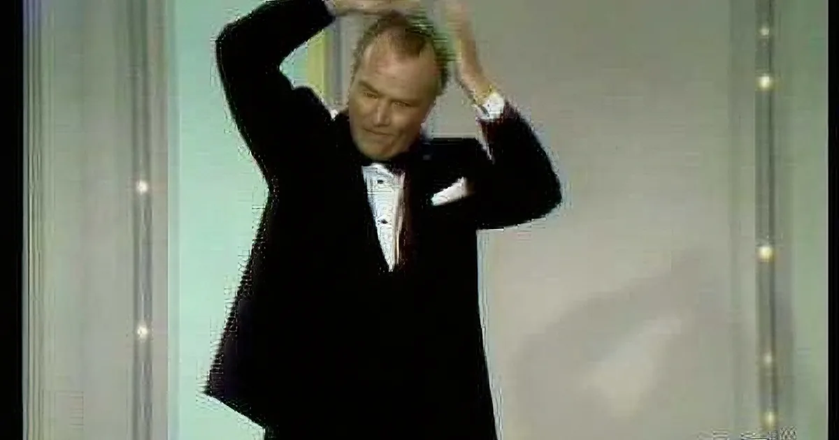 A man in a tuxedo adjusts his hair on stage, surrounded by a lighted backdrop.