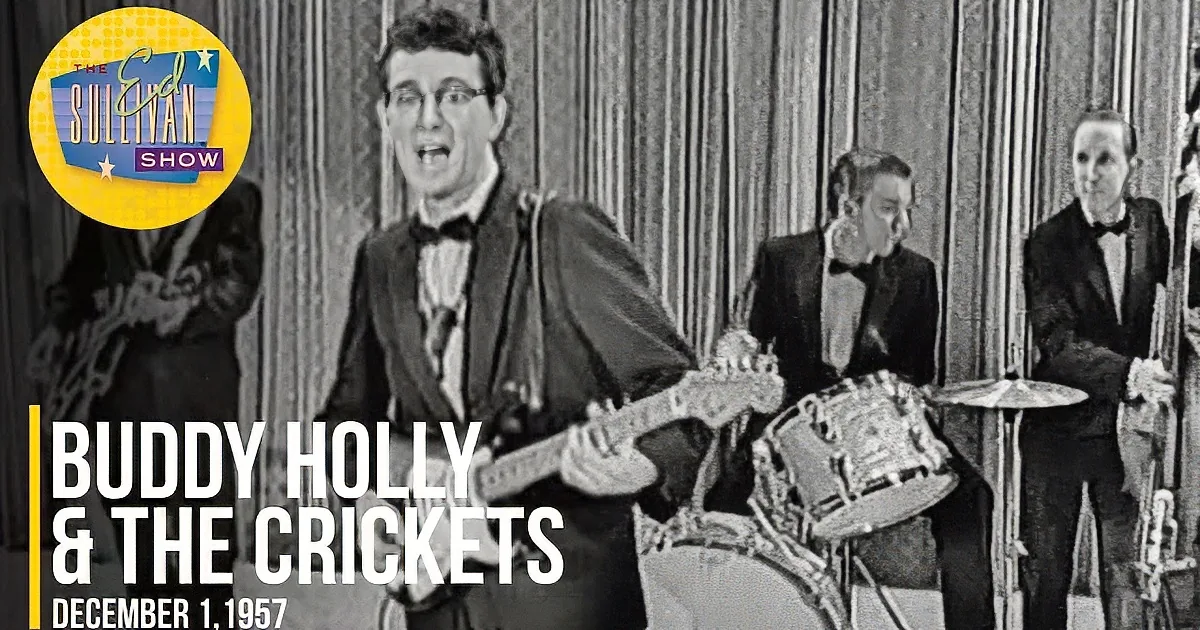 A man in a tuxedo plays an electric guitar, surrounded by a band in formal attire.