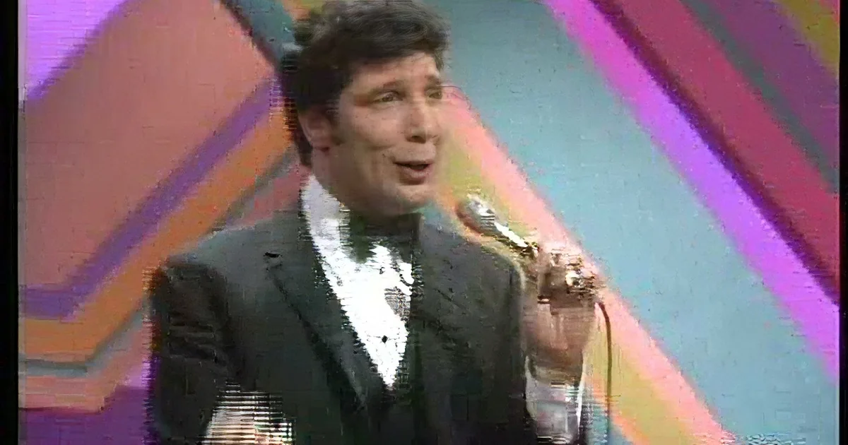 A man in a black suit and bow tie holds a microphone, singing on a stage with colorful geometric patterns.