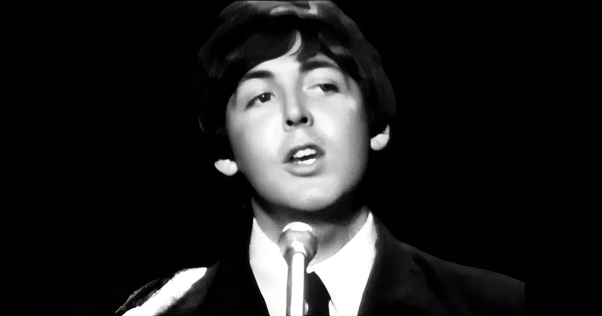 A man in a suit sings into a microphone against a dark background.