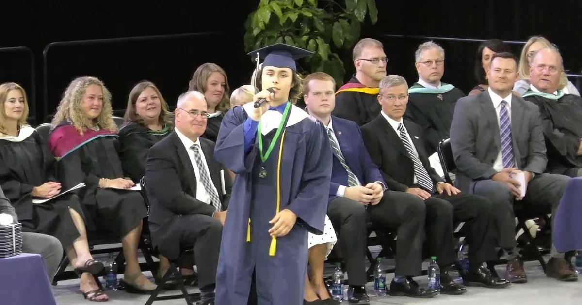 A graduate in a cap and gown holds a microphone, addressing an audience at a graduation ceremony.
