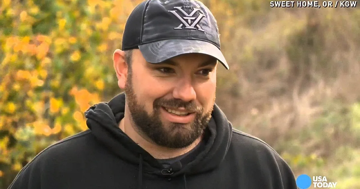 A man wearing a black hoodie and a camouflage baseball cap is standing outdoors with autumn foliage in the background.