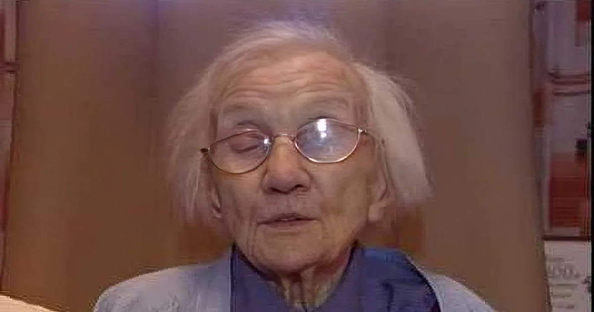 An elderly person with white hair and glasses, wearing a light blue shirt, is seated indoors.