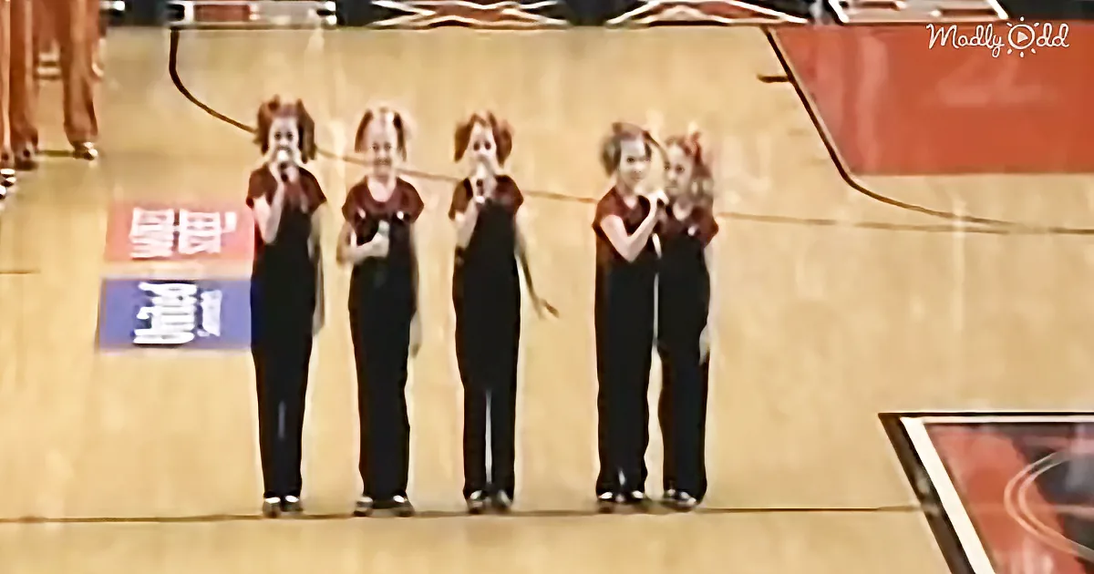 Four individuals in black outfits are standing in a row, holding microphones and singing on a basketball court.