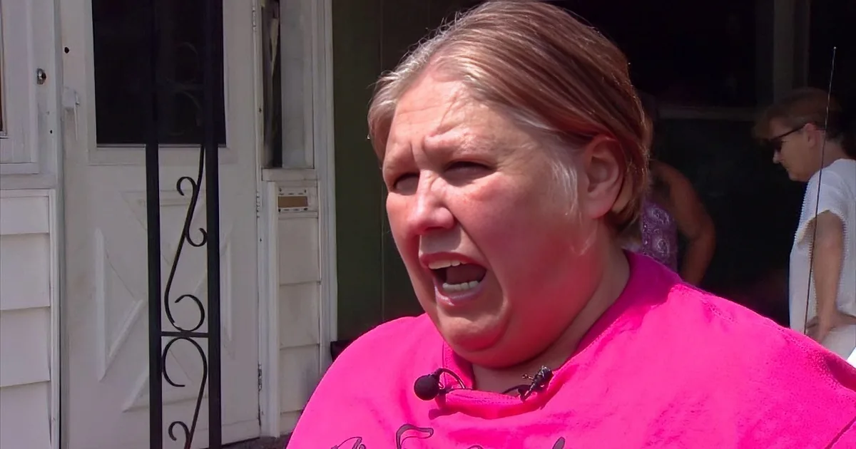 A woman in a bright pink shirt stands outside a house, speaking into a microphone.