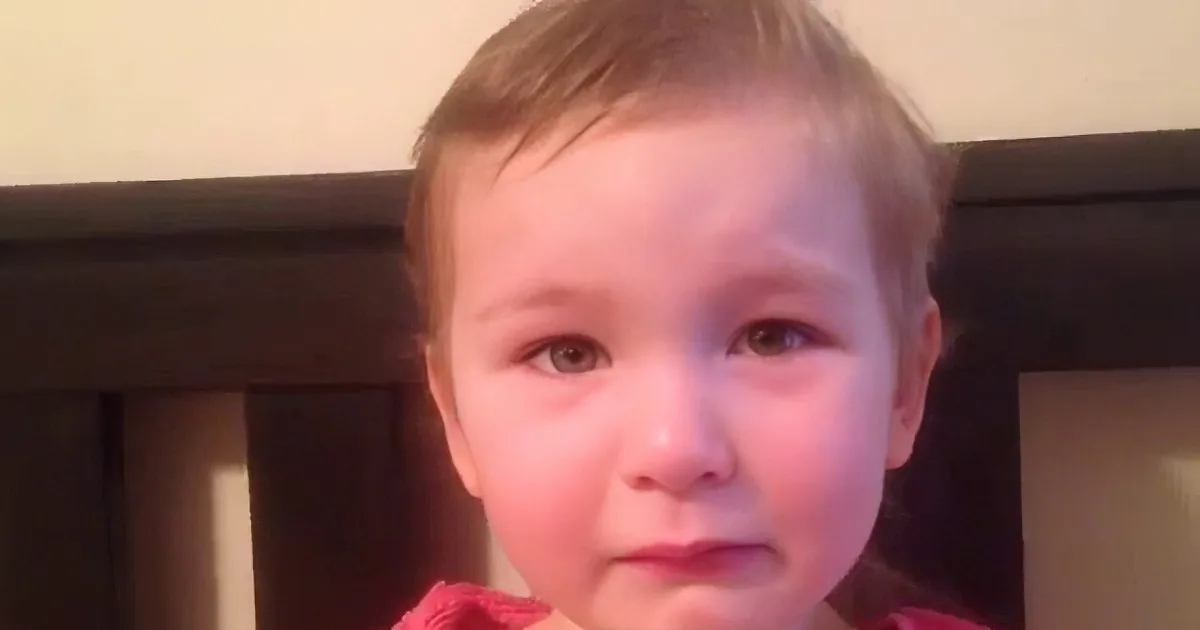 A young child with short hair, wearing a pink shirt, is looking directly at the camera.