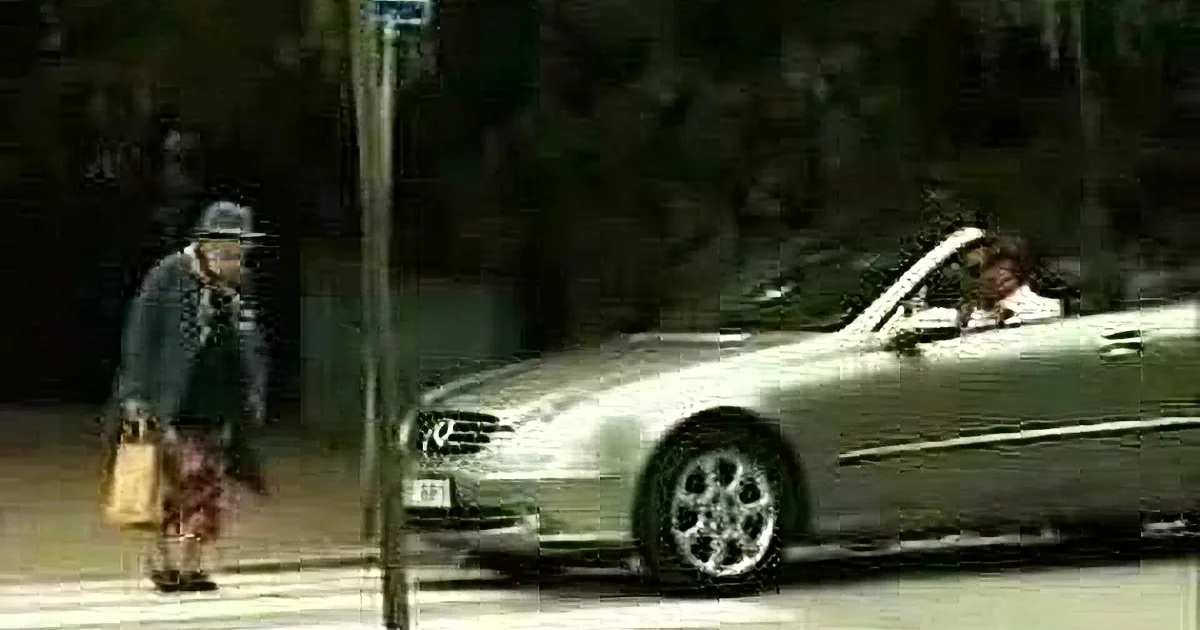 A man in a hat and coat walks past a silver car parked on a street.