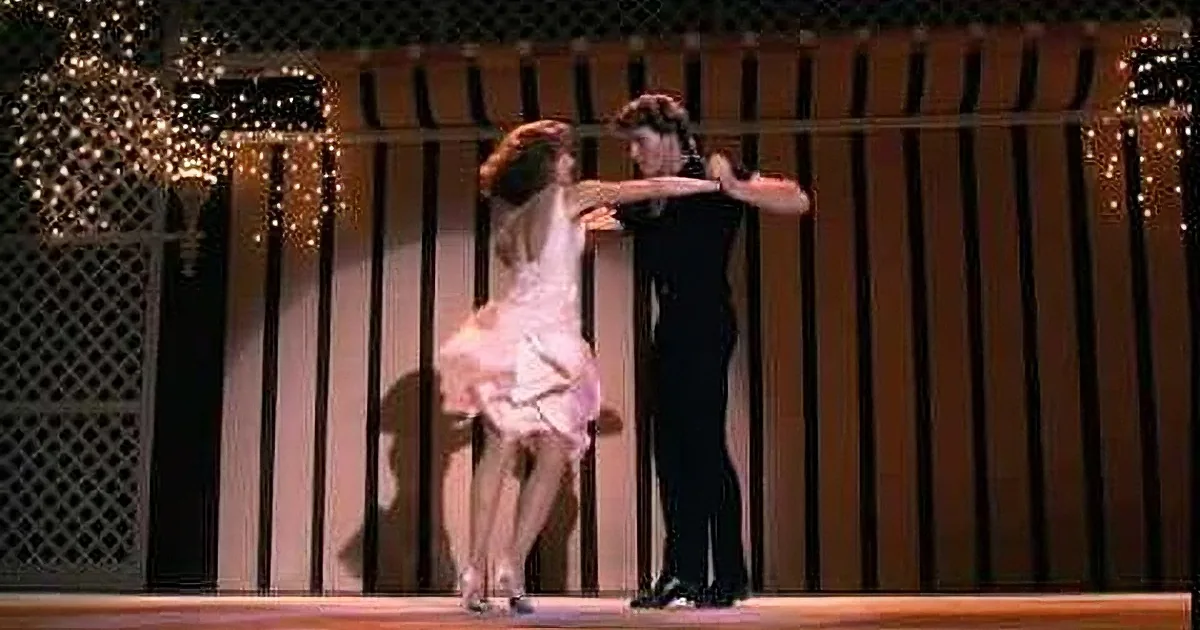 A couple dances on stage, woman in white dress, man in black suit, against a backdrop of vertical black and white stripes.