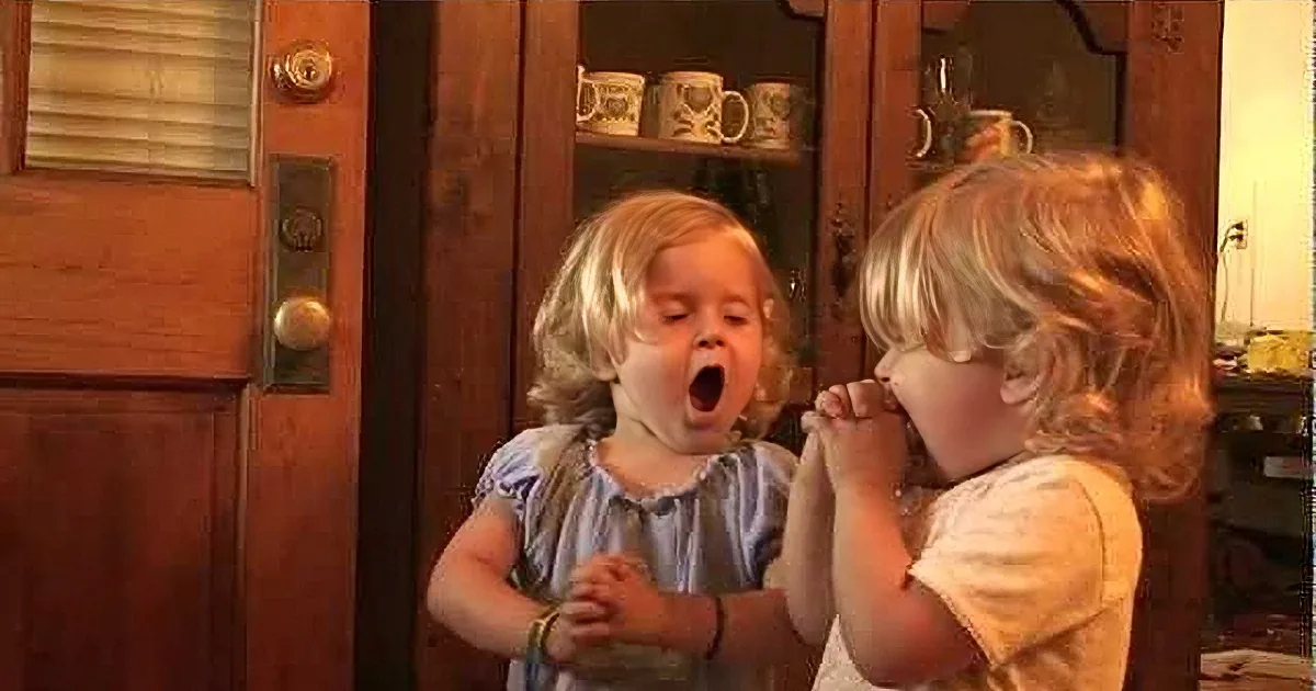 Two toddlers, one holding a spoon, the other crying, in a warmly lit room.