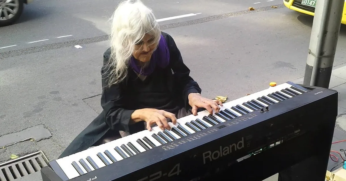 An elderly woman with long white hair plays a Roland keyboard on a city street.
