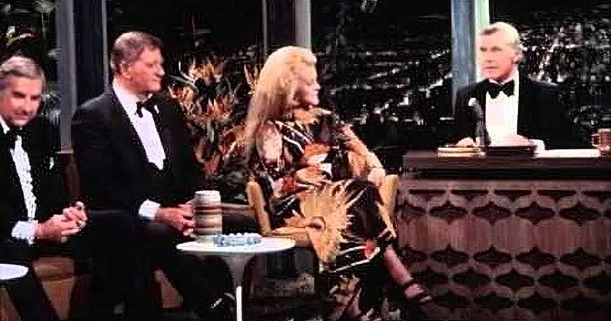 A woman in a leopard print dress sits between two men in tuxedos on a talk show set.