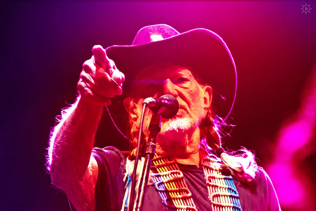 A man in a cowboy hat and fur vest sings into a microphone on stage with vibrant purple lighting.