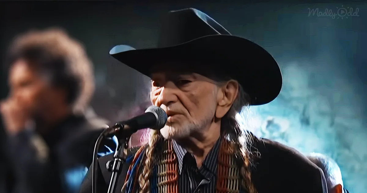 An older man wearing a black cowboy hat sings into a microphone, with another person in the background.