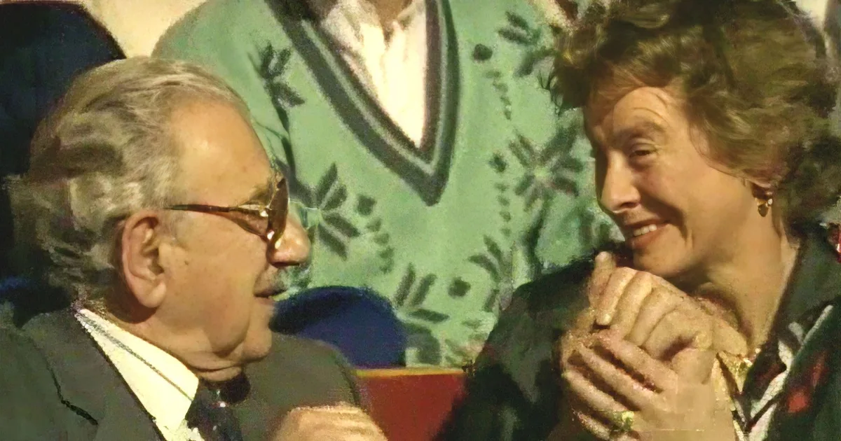 An older man in glasses and a woman with clasped hands smile at each other, possibly in a social setting.