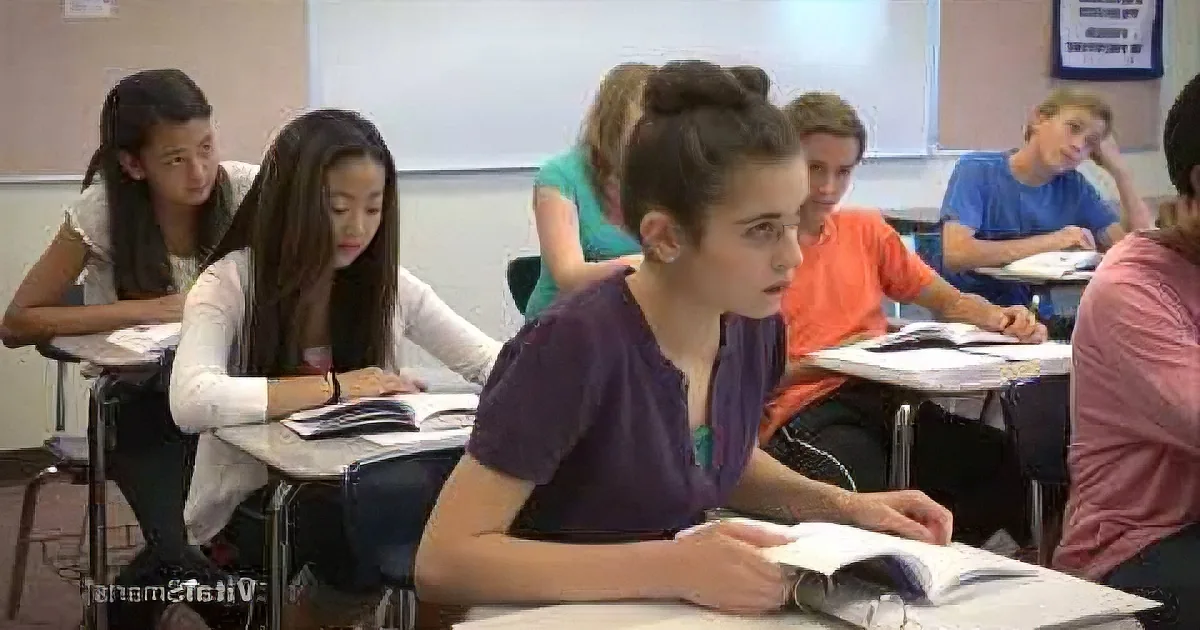A student in a purple shirt sits at a desk, reading a book in a classroom with other students.