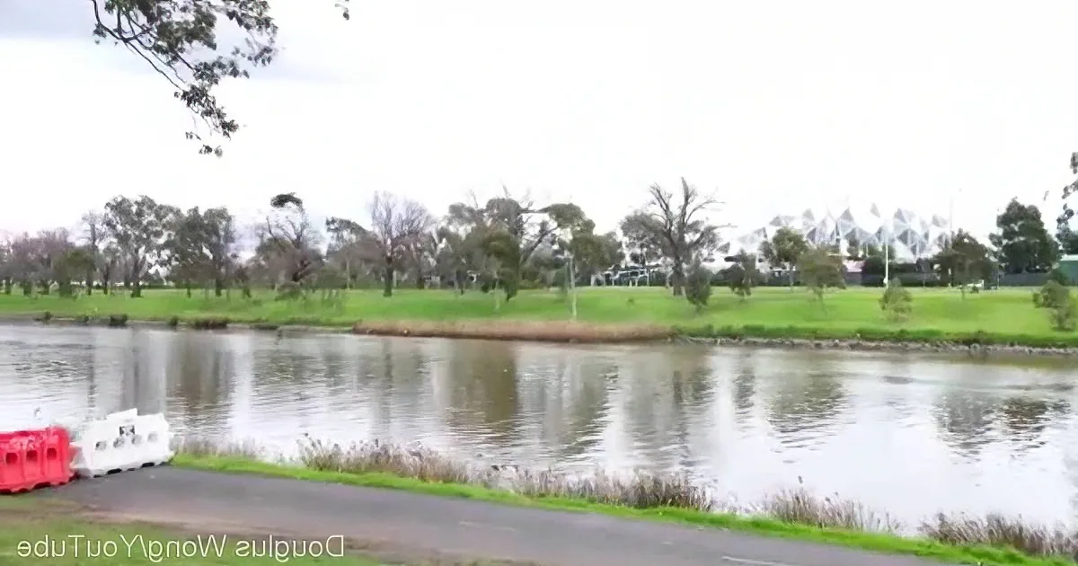 A serene river flows past a grassy bank with trees and a distant white structure.