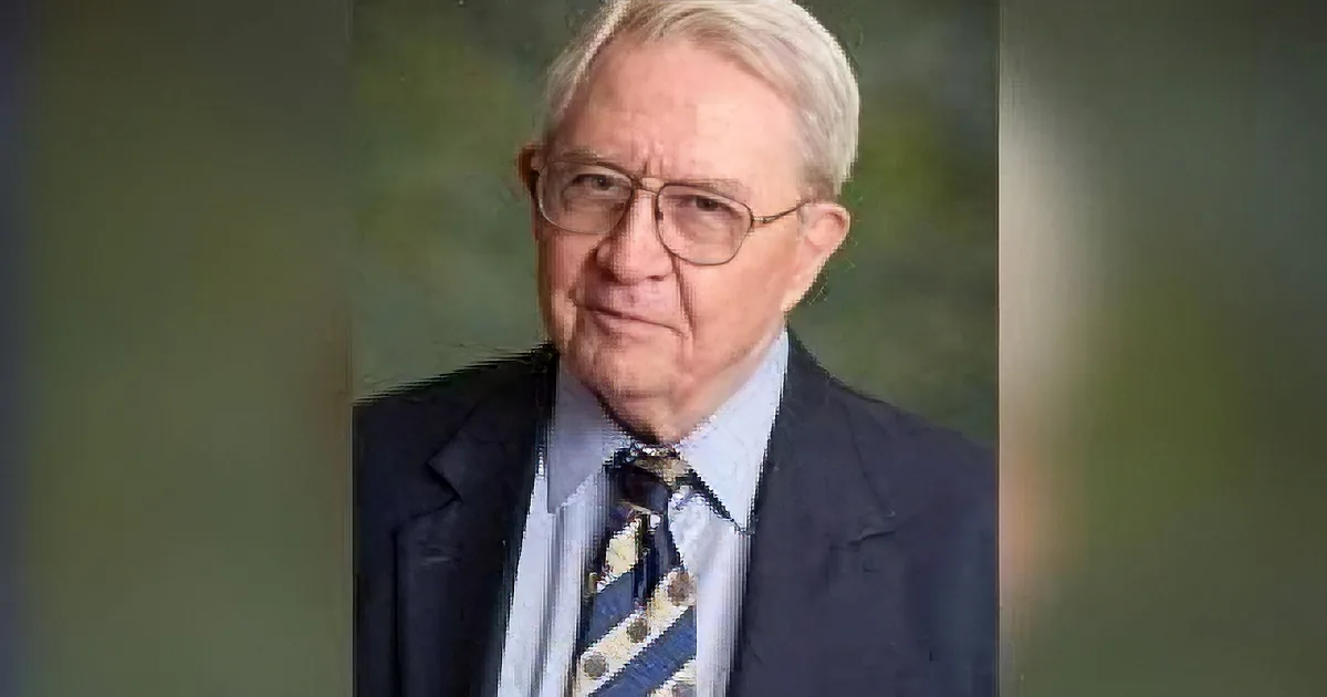 An older man in a suit and tie, wearing glasses, looks downward with a serious expression.