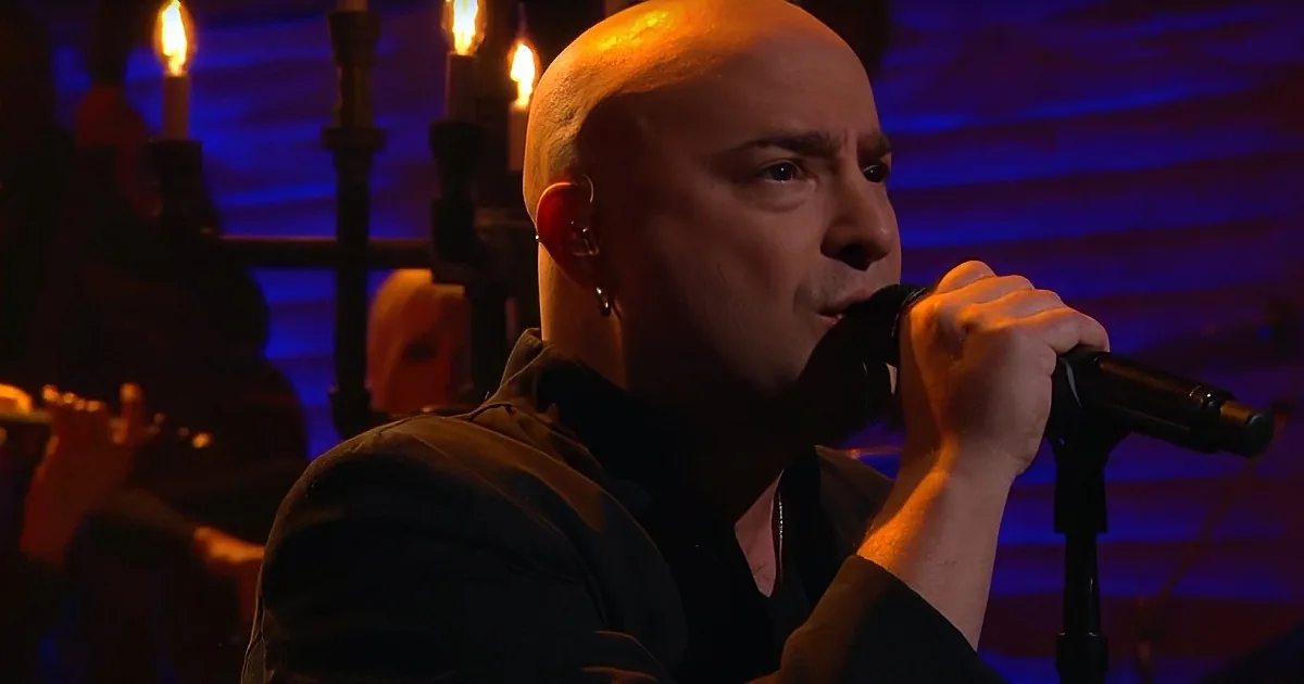 A person with a shaved head and ear piercings sings into a microphone in a dimly lit setting with candles.