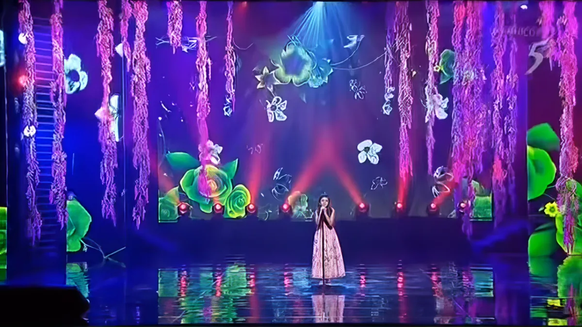 A performer stands on a vibrant, colorful stage with floral and light effects.