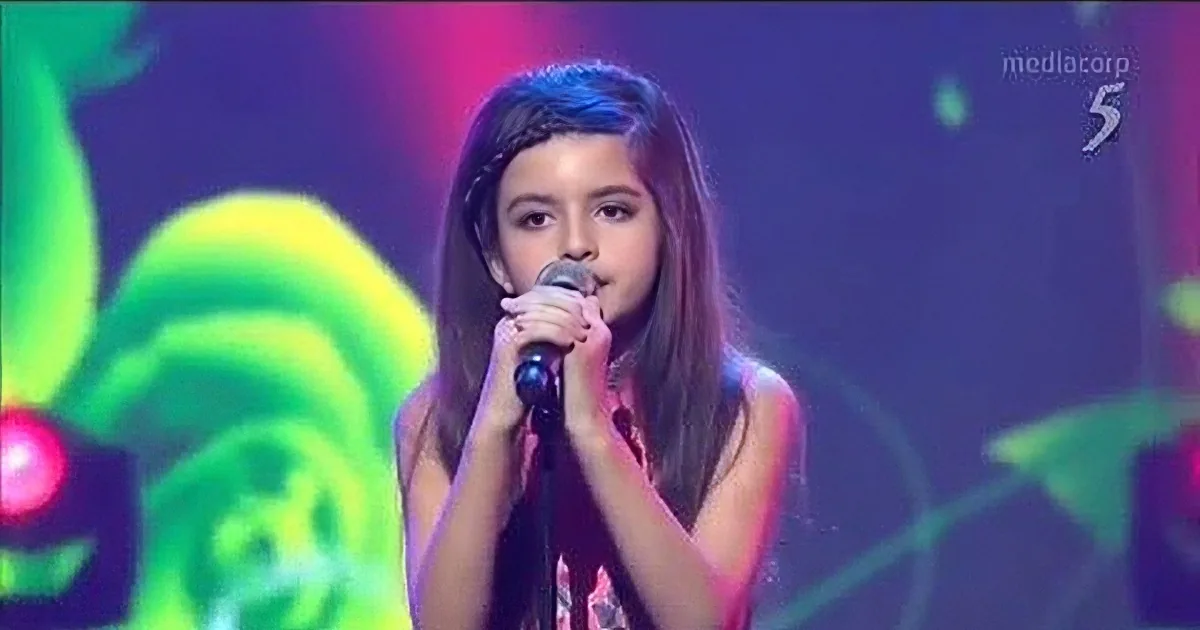 A person with braided hair sings into a microphone on a stage with vibrant red and purple lighting.