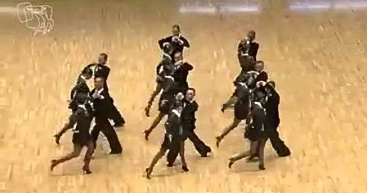 A group of people in dark uniforms is performing a synchronized dance routine on a sandy surface.