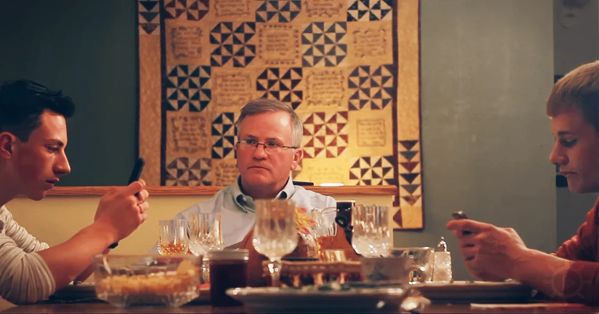 A man sits at a table with two others, holding phones, surrounded by glasses and food, with a quilted wall hanging in the...
