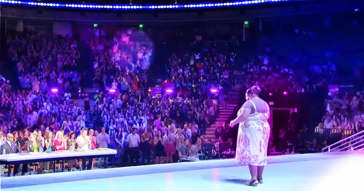 A woman in a white dress stands on a stage in front of a large, cheering audience in a brightly lit arena.