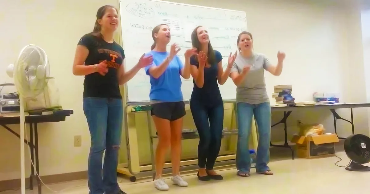 Four women in casual attire clap and sing in front of a whiteboard in a classroom setting.