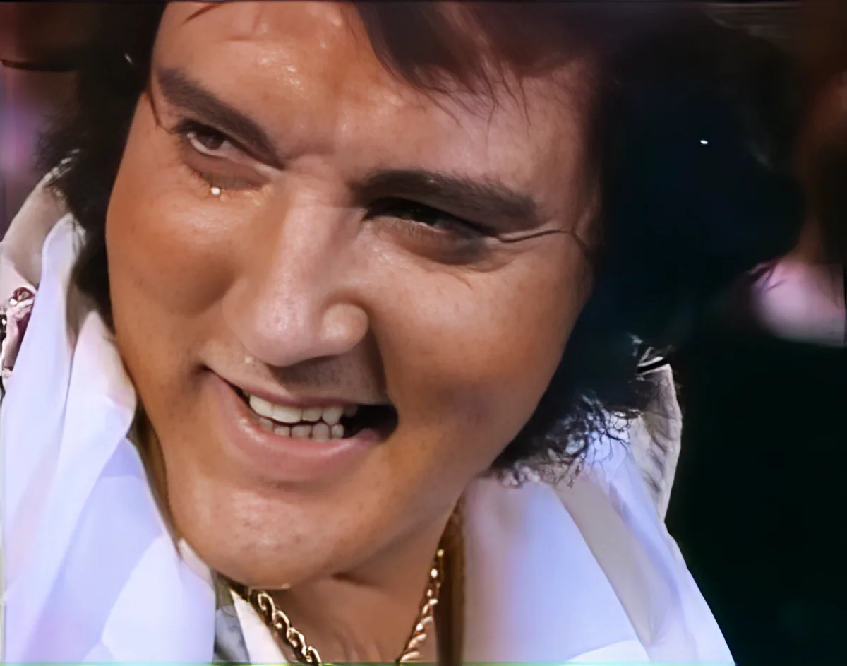 A person with dark hair and a white shirt is smiling, wearing a gold chain.