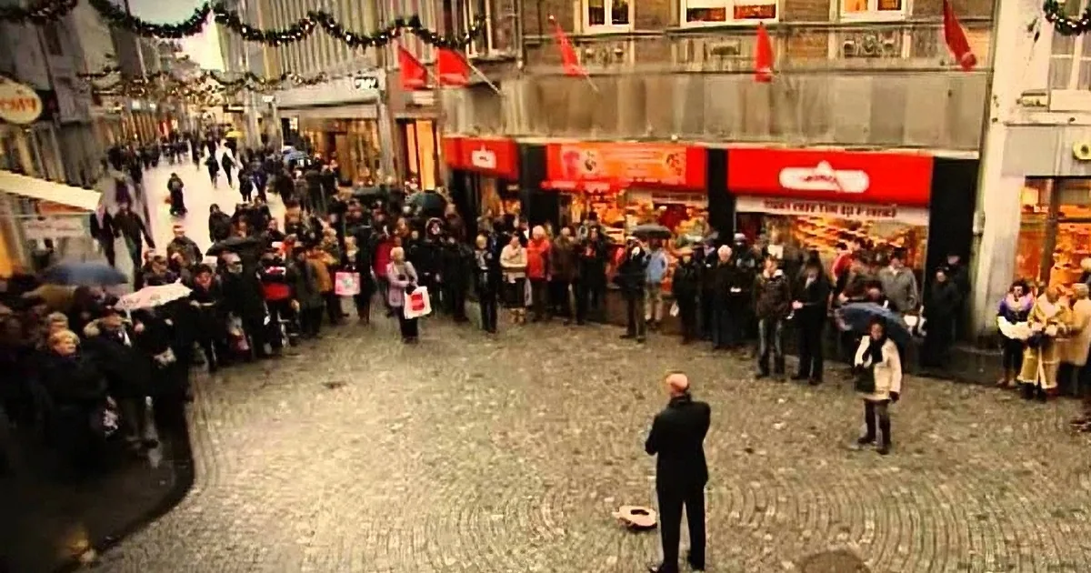 A crowd gathers in a cobblestone square, some holding signs, under festive Christmas decorations.