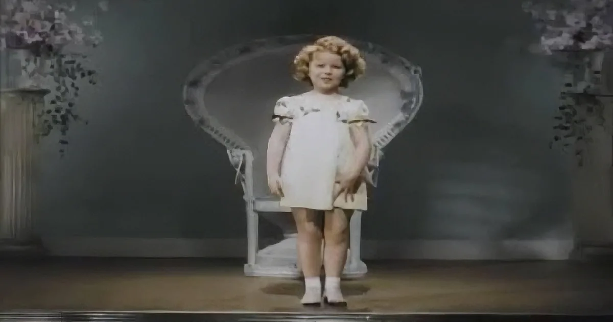 A young child in a white dress stands in front of a decorative chair, with floral decorations on either side.