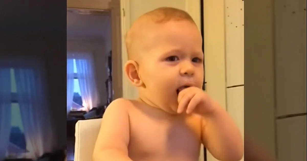 A baby with short hair, shirtless, holding a white object near its mouth.