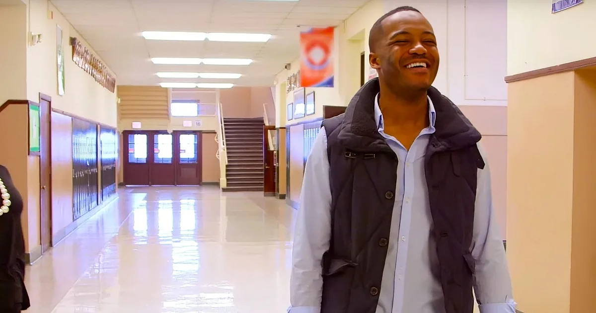 A man in a vest and shirt smiles in a school hallway with lockers and a staircase in the background.