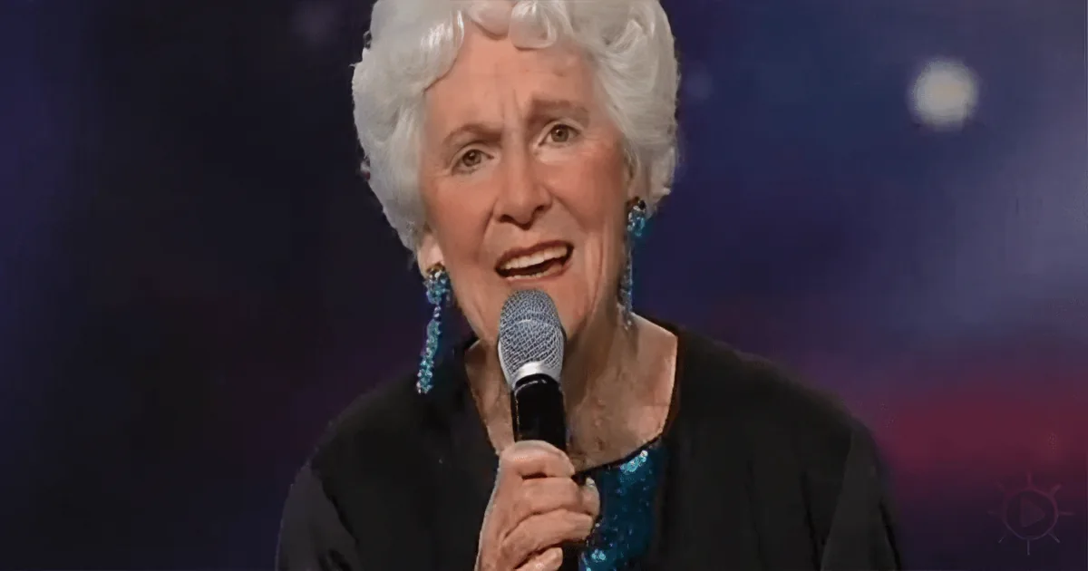 An elderly woman with white hair and blue earrings sings into a microphone on stage.