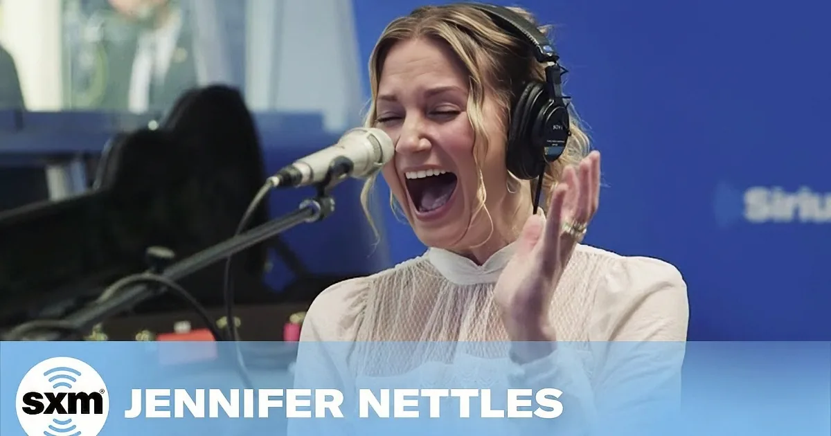A woman with blonde hair, wearing headphones, sings into a microphone in a studio setting.