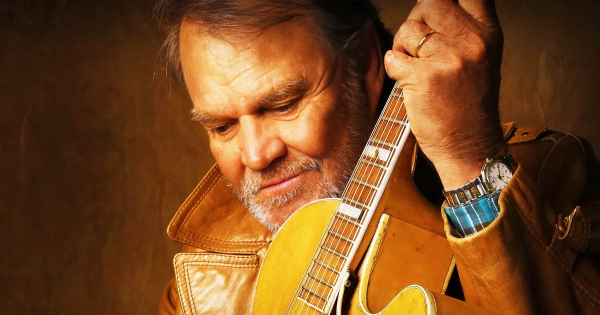 A man in a brown leather jacket holds a guitar, looking down with a thoughtful expression.