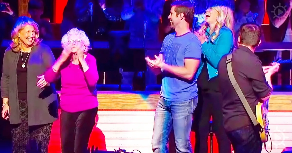 A group of people, including a woman in a pink shirt, are clapping and smiling on a stage with colorful lighting.