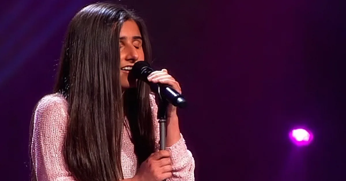 A woman with long dark hair sings into a microphone on a stage with purple lighting.