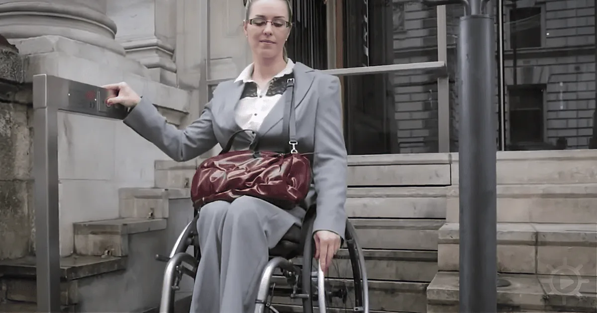A woman in a wheelchair, wearing a suit, uses a security turnstile outside a building.
