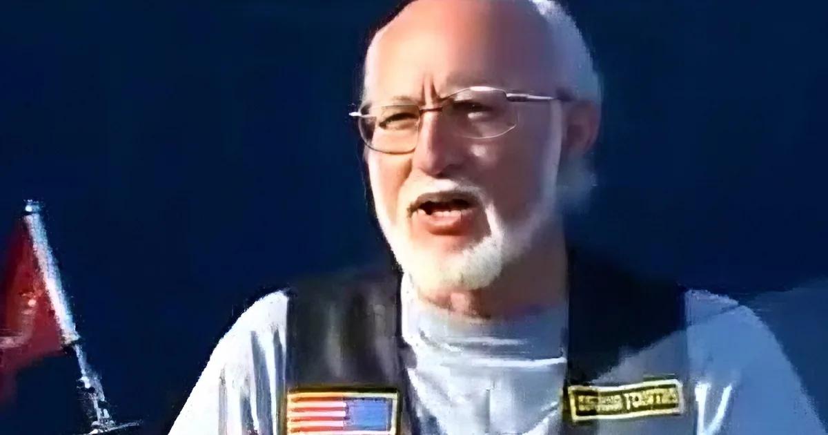 A man with a white beard and glasses holds an American flag patch, standing in front of a dark background with flags.