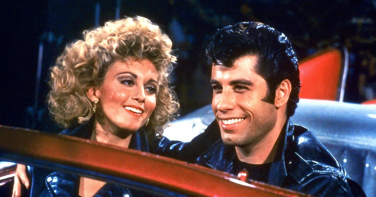 A man and woman in 1950s attire smile, the woman with blonde curls, the man with slicked-back hair.