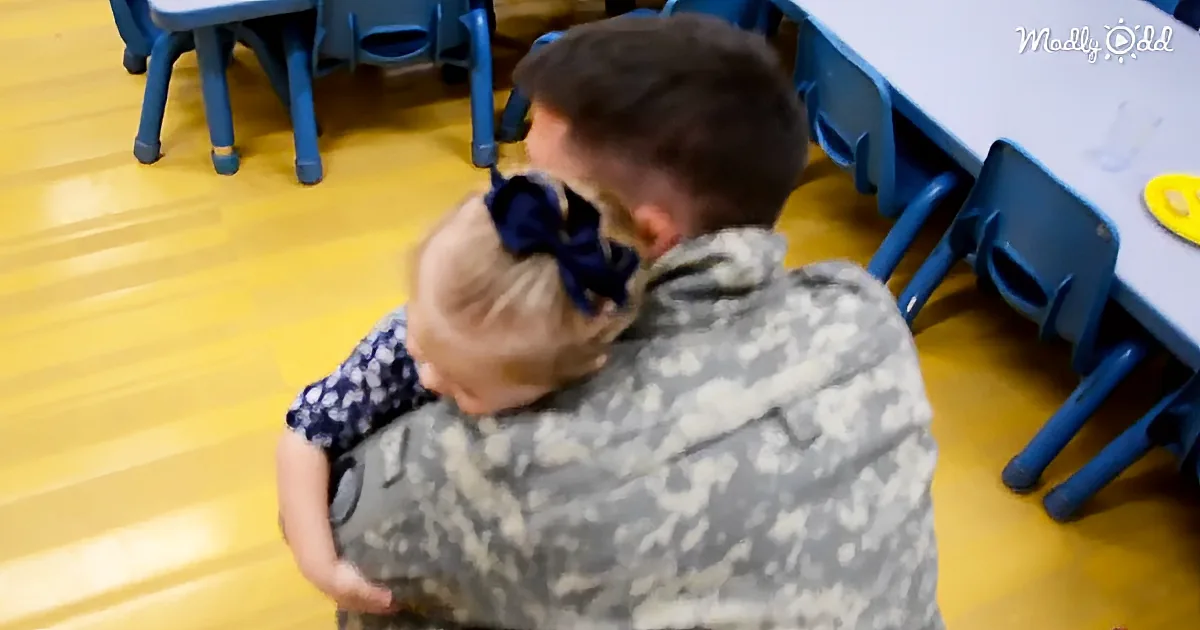A soldier in camouflage hugs a child with a blue bow in a classroom setting.